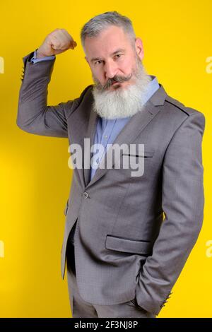 Studio portrait mature businessman dressed in gray suit shows his pumped up biceps, strong business, exercising in entrepreneurship concept. Stock Photo