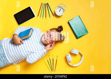 Schoolgirl little girl in glasses with a book lies on a yellow background. Top view of a child student surrounded by a tablet, pencils, alarm clock an Stock Photo