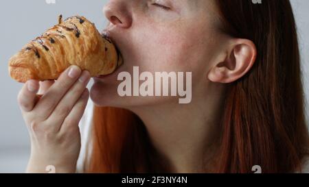 Happy young woman eats a croissant sitting on couch at home at morning Stock Photo