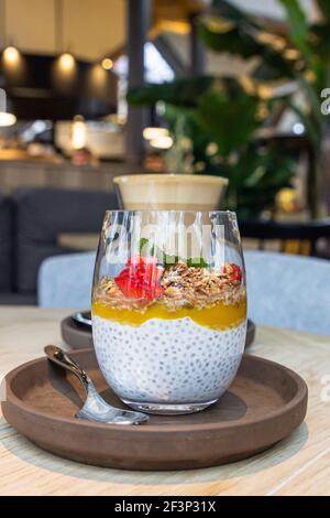 Chia pudding with mango puree, oats flakes and fresh strawberries and cup with cappuccino on wooden table in cafe. Selective focus. Stock Photo