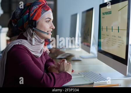 Young muslim woman in headphones looking at computer monitor and talking on the phone working in call center Stock Photo