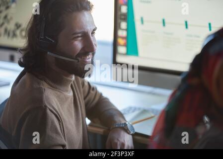 Young bearded man in headphones talking to his colleague while sitting at the table in front of computer Stock Photo