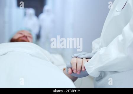 Close-up of doctor in protective gloves holding hands of the patient and supporting him while he lying on gurney Stock Photo