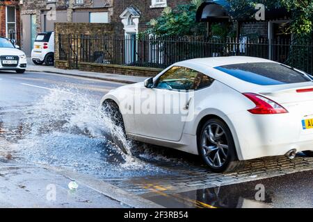 A car drives through a large roadside puddle caused by a mains water leak, showering the pavement with spray, London, UK Stock Photo