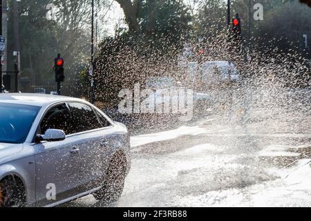 A car drives through a large roadside puddle caused by a mains water leak, showering the pavement with spray, London, UK Stock Photo