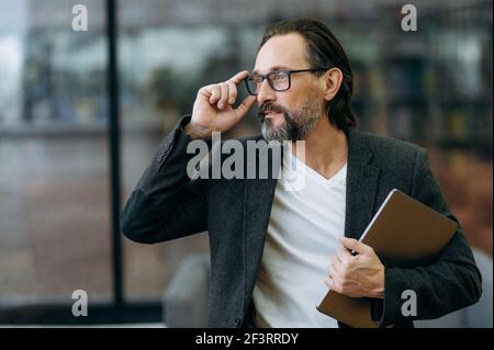 Middle-aged caucasian successful man in the office. A stylish handsome senior businessman in a formal wear with a beard holds a laptop in his hand and looks away, adjusting his own glasses, smiling Stock Photo