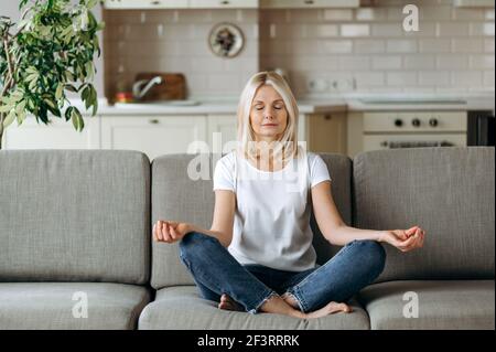 Calm adult lady is resting at home in the living room. Beautiful middle-aged caucasian blonde in stylish casual wear is sitting on the sofa, meditating with her eyes closed in lotus position, chilling Stock Photo