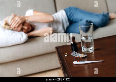 Defocused sick adult woman at home on the couch. A middle-aged caucasian ill woman have headache, clutching her head, a glass of water on the table and a thermometer with pills, need a treatment Stock Photo