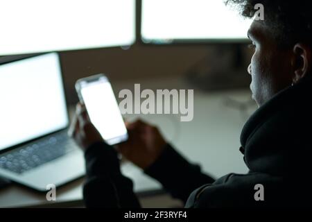 Close up shot of busy young guy sitting in dark room in front of many computer monitors and using smartphone while working alone late at night Stock Photo