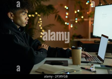 Side view of stylish young guy looking at monitor screen while working on laptop computer at late night Stock Photo
