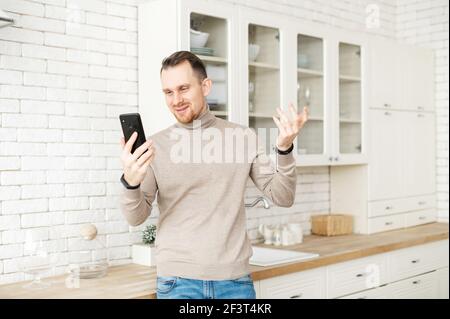 Cheerful guy using smartphone for video call staying at home. Smiling handsome man holds a phone and waving hello into selfie camera, has virtual meeting with a family, colleagues Stock Photo