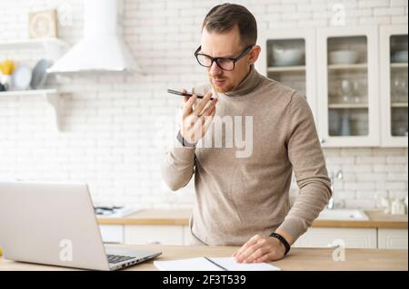 Modern young businessman wearing stylish eyeglasses and turtleneck working remotely at home, a guy is recording voice message, looking through workpapers and making voice notes, using laptop Stock Photo