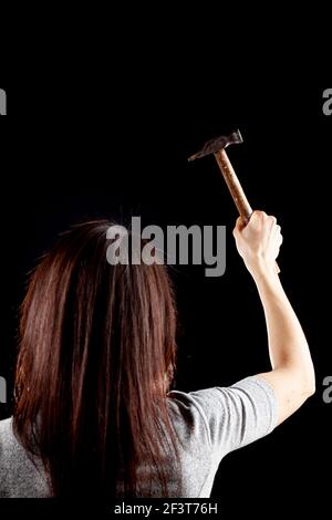 A rim light dark background image of a young caucasian woman with long brown hair. She is raising her right hand holding a hammer. A protest, activism Stock Photo