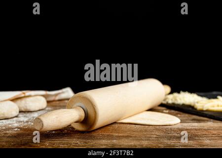 An American wooden rolling pin on  a flattened dough on wood kitchen countertop. A bread making concept image with loafs and cheese toppings in blurre Stock Photo