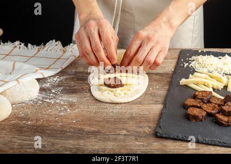 a woman is placing ingredients on flattened dough on wood kitchen countertop. A bread making concept image with loafs,  cheese and sucuk in back. Turk Stock Photo