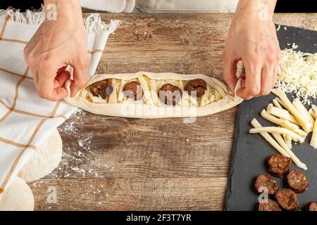 a woman is placing ingredients on flattened dough on wood kitchen countertop. A bread making concept image with loafs,  cheese and sucuk in back. Turk Stock Photo