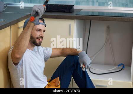 Happy worker. Portrait of cheerful young handyman holding a hammer and an adjustable wrench in his hands while sitting on the floor in the kitchen Stock Photo
