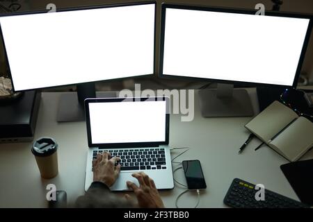Work anytime. High angle view of hands of man sitting at the table in front of many computer monitors and using laptop while working late at night Stock Photo