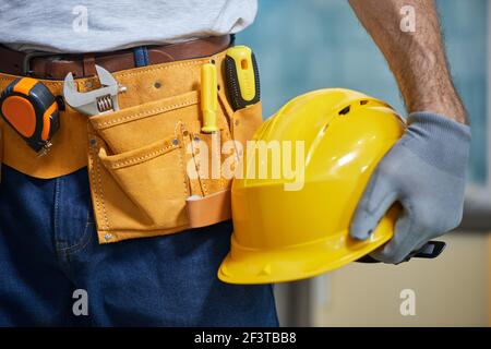 Close up of repairman wearing a tool belt with various tools, holding a helmet in his hand Stock Photo