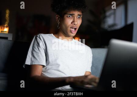Amazement. Night portrait of emotional young guy looking surprised, surfing the internet, using laptop in dark room, sitting on a couch at home Stock Photo