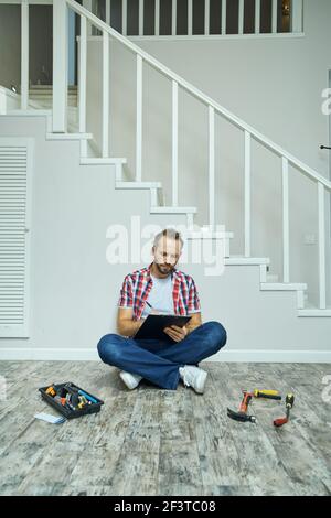 Busy handyman making notes on clipboard while sitting on the floor in apartment. Home repair hand tools on laminate flooring Stock Photo