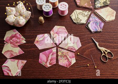 Pentagonal pieces of fabric lying in the shape of a flower, stacks of multi-colored pieces of fabric, sewing accessories on a desk