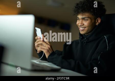 Distraction. Portrait of happy male student smiling and using smartphone while studying, working on laptop late at night Stock Photo