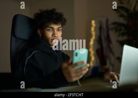 Stylish young guy with piercing looking at smartphone screen while sitting at the table, using laptop and working late in the evening Stock Photo