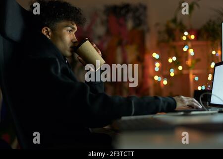 Coffee break. Side view of young guy with piercing drinking coffee, sitting at the table in front of many computer monitors and working from home late Stock Photo