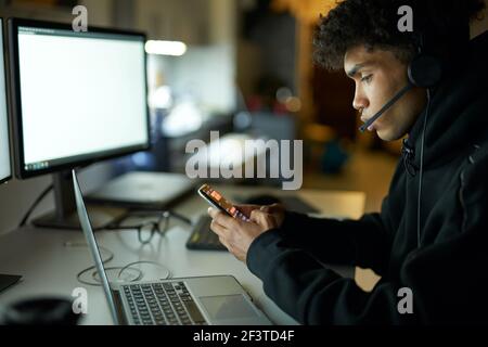 Side view of young guy wearing headset sitting at the table in front of computer monitors and texting using smartphone while working from home in dark Stock Photo