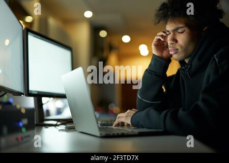 Exhaustion. Tired young guy having headache after working too long on laptop, sitting with eyes closed in dark room at home late at night Stock Photo