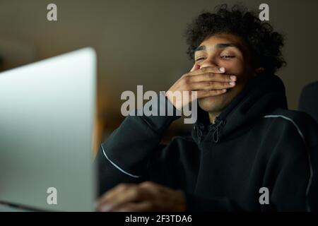 Tiredness. Sleepy young guy holding hand over his mouth while yawning. Male student studying using laptop, sitting in dark room at home late at night Stock Photo
