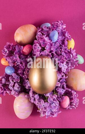 Easter festive concept with big decorative gold egg on a flowers of lilac against bold pink background. Spring festive backdrop. Stock Photo
