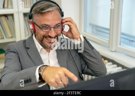 Smiling Mature Businessman With Headphones Sitting at Office Desk Pointing at Computer Monitor. Excited Businessman Gesturing with Hand in His Office. Stock Photo