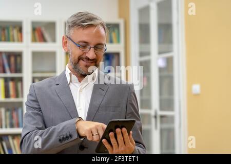 Handsome Mature Businessman or Teacher Standing in front of Bookshelves and Holding Tablet Computer. Business Executive Using Tablet PC. Stock Photo