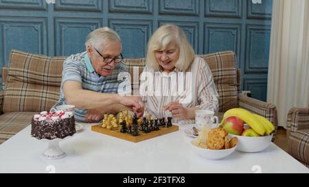 Retired senior couple talking drinking tea, playing chess in modern living home room lounge together Stock Photo