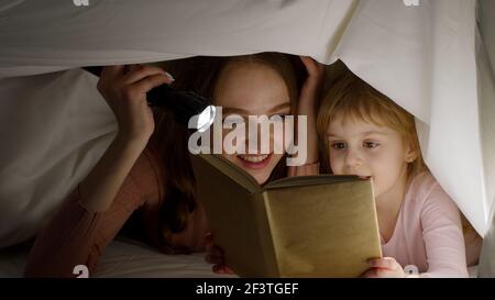 Young mother reading goodnight story fairytale to child daughter under duvet blanket in night room Stock Photo