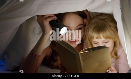 Young mother reading goodnight story fairytale to child daughter under duvet blanket in night room Stock Photo