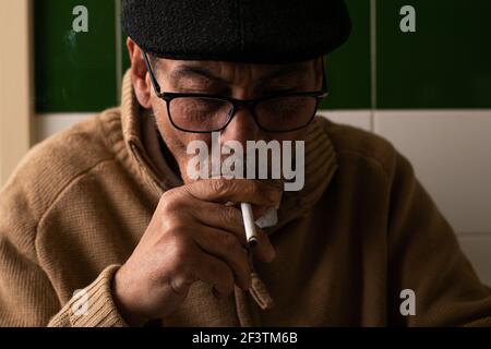 Close-up of an elderly Caucasian man, wearing a beige sweater, dark gray beret and black glasses, holding a smoking cigarette to his mouth in a though Stock Photo