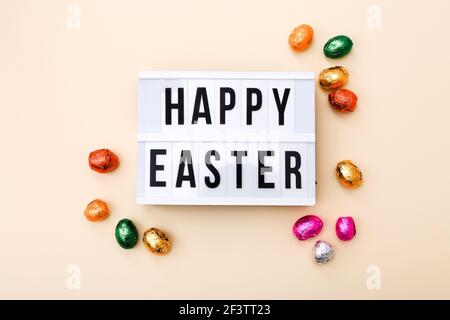 Easter wishes on lightbox with chocolate mini eggs wrapped in a colorful foil on a beige background, top view Stock Photo