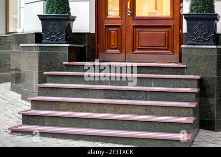 marble staircase with granite steps to wooden entrance door on store with handle and glass, threshold and pedestals for flowerpots with bushes at faca Stock Photo