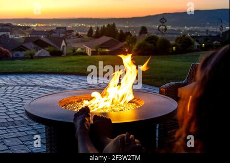 A woman relaxes by a roaring firepit on a paver patio at sunset overlooking the Spokane Valley, in Washington State, USA Stock Photo