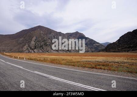 A two-lane asphalt road with fences, leading directly through a picturesque autumn valley at the foot of high mountains. Chuisky tract, Altai, Siberia Stock Photo