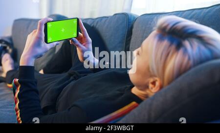 Woman lying on a couch using smartphone with green mock-up screen. High quality photo Stock Photo