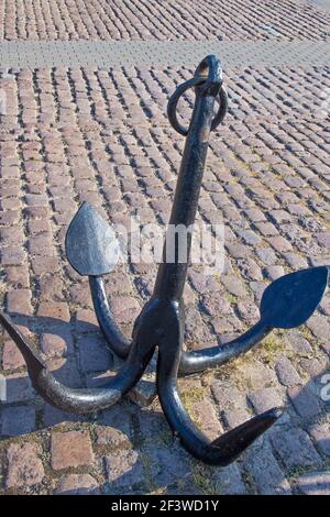 An old four legged anchor with almond shaped points at the ends painted black with a ring at the top standing on a paved square of rough brown paving Stock Photo