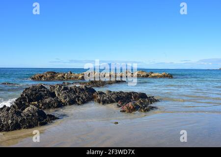 Flat formation of dark rocks exposed on sandy beach during low tide with island far in background. Stock Photo