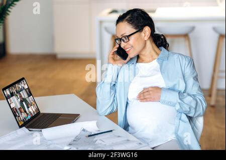 Successful busy mixed race pregnant adult woman is sitting near her work desk, having pleasant cellphone business conversation and in parallel an online conference with colleagues on a laptop, smiling Stock Photo