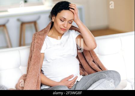 Tired sad pregnant mixed race adult woman feeling bad and unhealthy, cold, flu, sitting on sofa in living room under plaid with closed eyes, need a rest and treatment Stock Photo