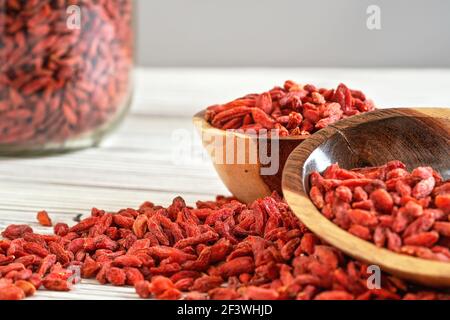 Dried goji berries in wooden bowls, scattered over white boards table under, blurred large glass jar full of fruits in background Stock Photo