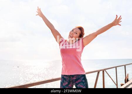 An adult woman in sportswear smiles happily and poses with her hands raised in the air against the sky and sea. Copy space. The concept of sport and a Stock Photo